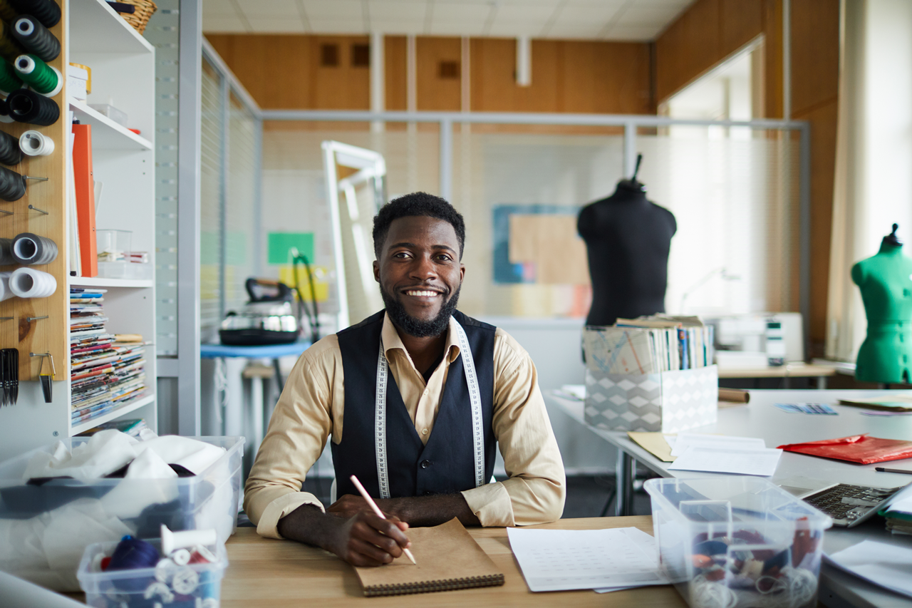 photo of smiling man in yellow shirt and sewing studio writing in notebook